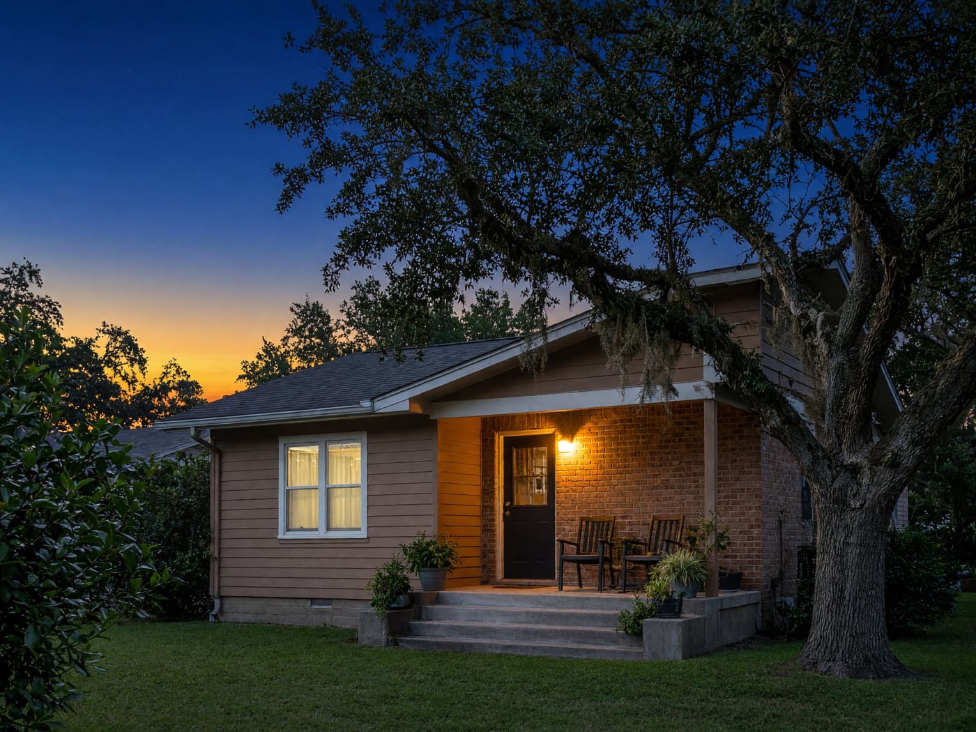 The front porch of a small brick and siding house at dawn, with the porch light on and a live oak overhead.
